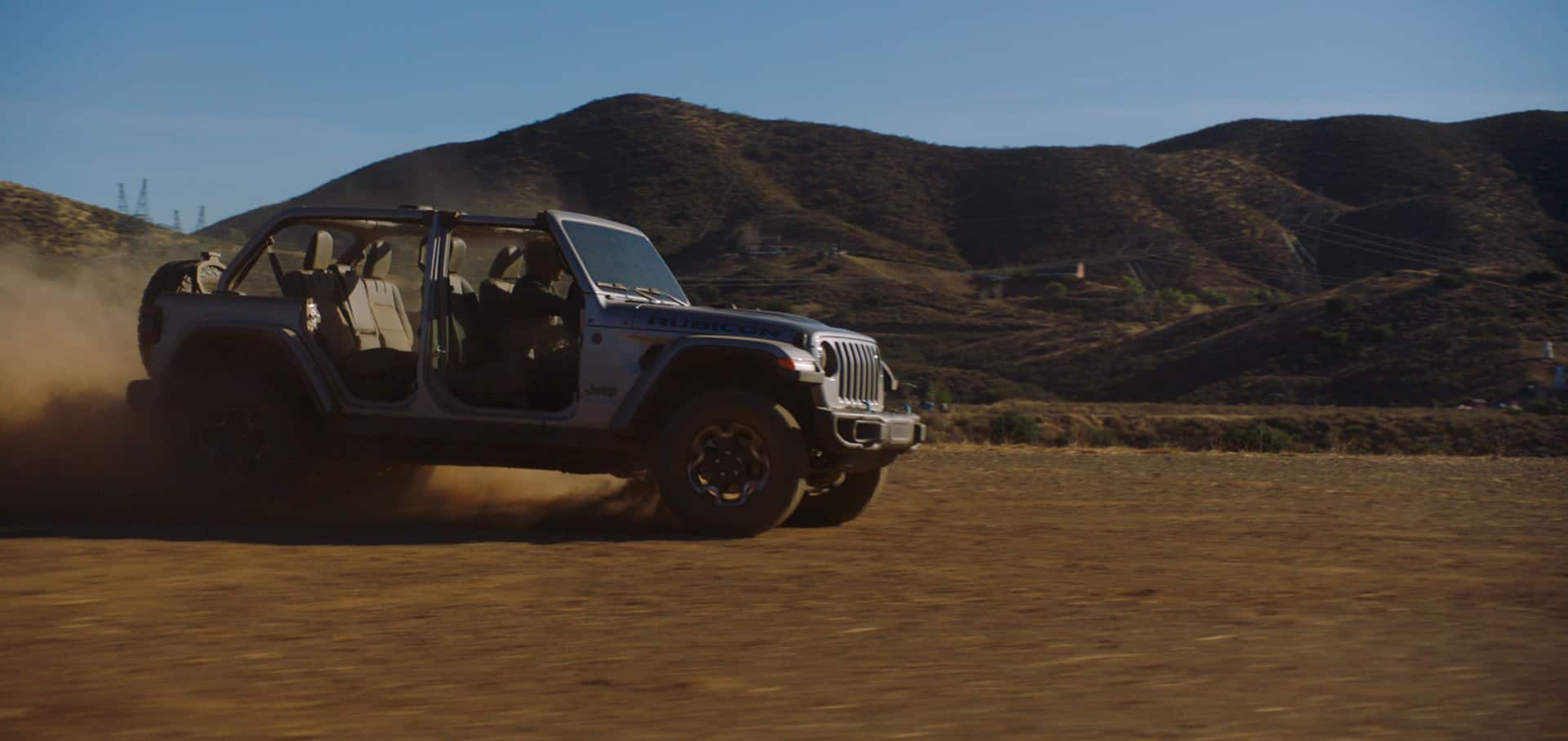 The 2023 Jeep Wrangler Rubicon 4xe being driven on sand with its doors off and a cloud of dust obscuring its rear wheels.