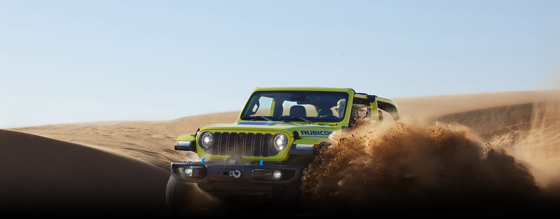 A lime green 2024 Jeep Wrangler Rubicon 4xe climbing a dune, kicking up clouds of sand as it goes.